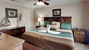 A neatly arranged bedroom with a large bed, a bedside table, a nightstand, a desk, and a ceiling fan. - Just for Fun by Ryson Vacation Rentals (Galveston)