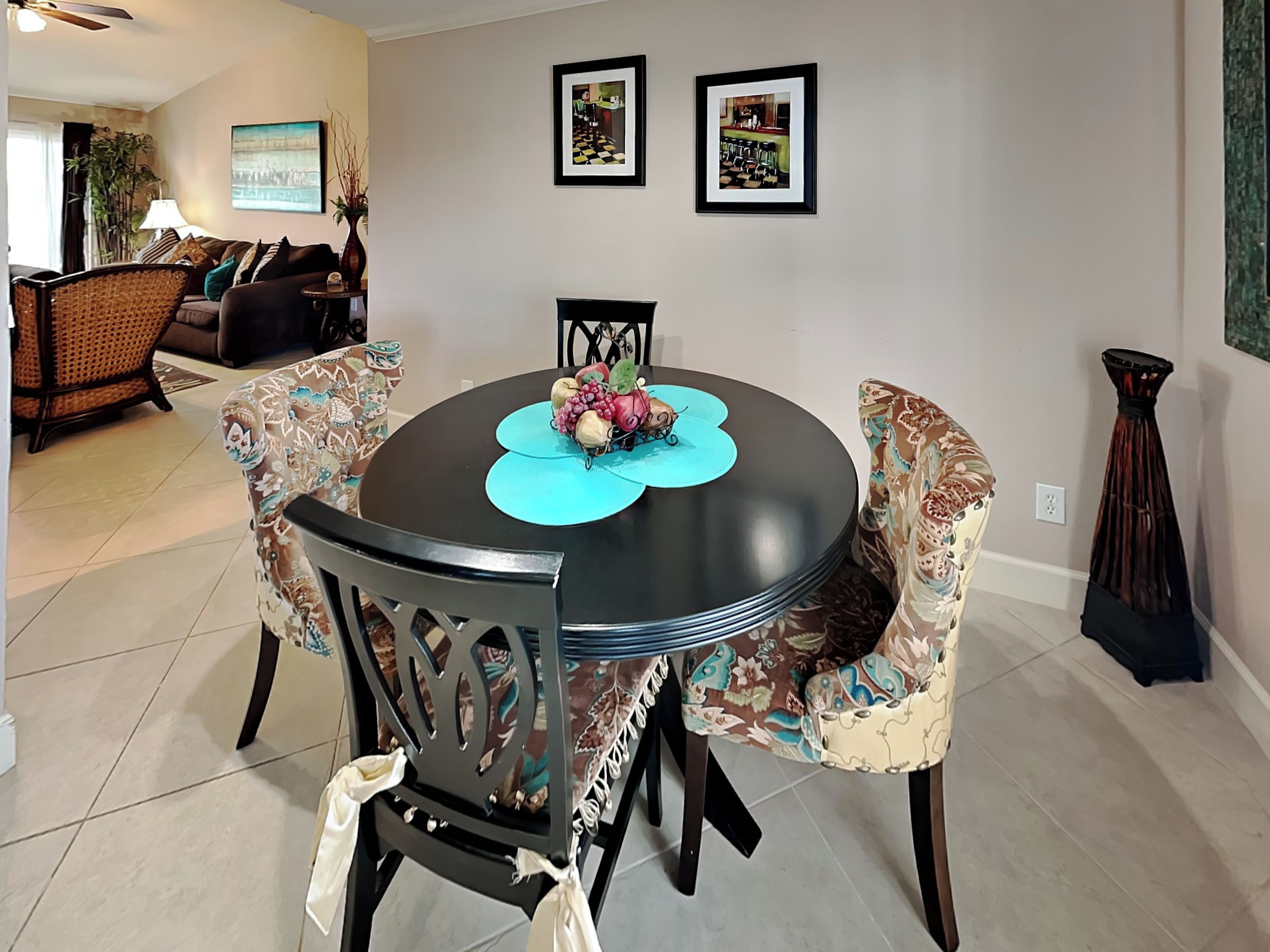 A dining area with a round table, floral-patterned chairs, and a vase of flowers. There are two framed pictures on the wall and a tall, narrow vase on the right.