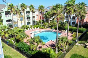A pool surrounded by palm trees and a multi-story building with balconies. - Just for Fun by Ryson Vacation Rentals (Galveston)
