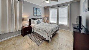 Un dormitorio con cama, mesitas de noche, escritorio, ventilador de techo y ventana con persianas.