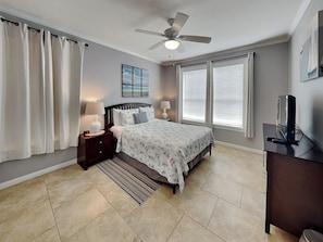 A bedroom with a bed, nightstands, a desk, a ceiling fan, and a window with blinds.