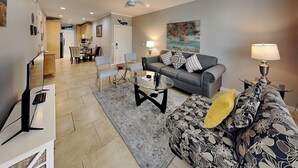 Una sala de estar con un sofá gris, un reposapiés estampado, una mesa de centro de vidrio y un mueble para televisión.