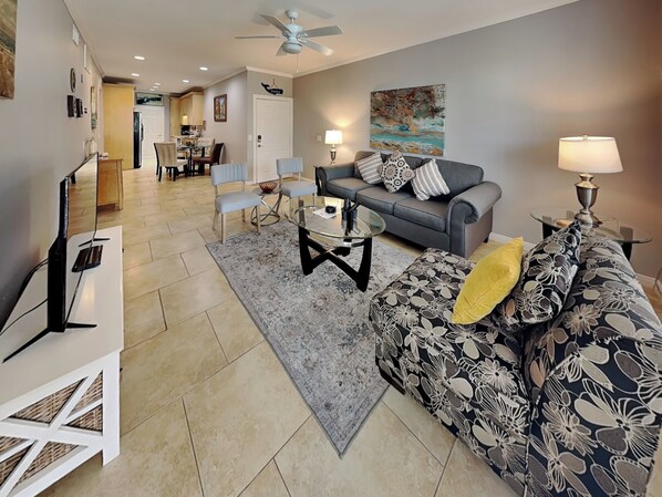 A living room with a grey sofa, a patterned ottoman, a glass coffee table, and a TV stand.