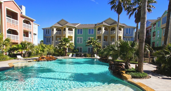 A swimming pool surrounded by multi-story buildings with balconies, palm trees, and a clear blue sky.