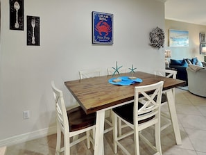 A dining area with a wooden table and white chairs, a poster on the wall, and a view of the living room.