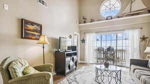 A living room with a wicker chair, a glass coffee table, a TV, and a balcony with a view. - The Regal Seagull by Ryson Vacation Rentals (Galveston)