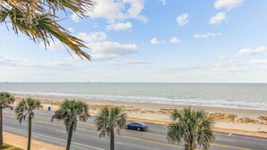 A coastal road with palm trees and a beach in the background. - The Regal Seagull by Ryson Vacation Rentals (Galveston)