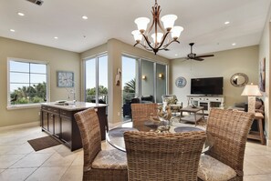 A spacious living area with a large dining table, wicker chairs, a chandelier, and a television. - Sapphire by Ryson Vacation Rentals (Galveston)