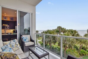 A balcony with a glass table and wicker furniture, overlooking a beach and palm trees. - Sapphire by Ryson Vacation Rentals (Galveston)