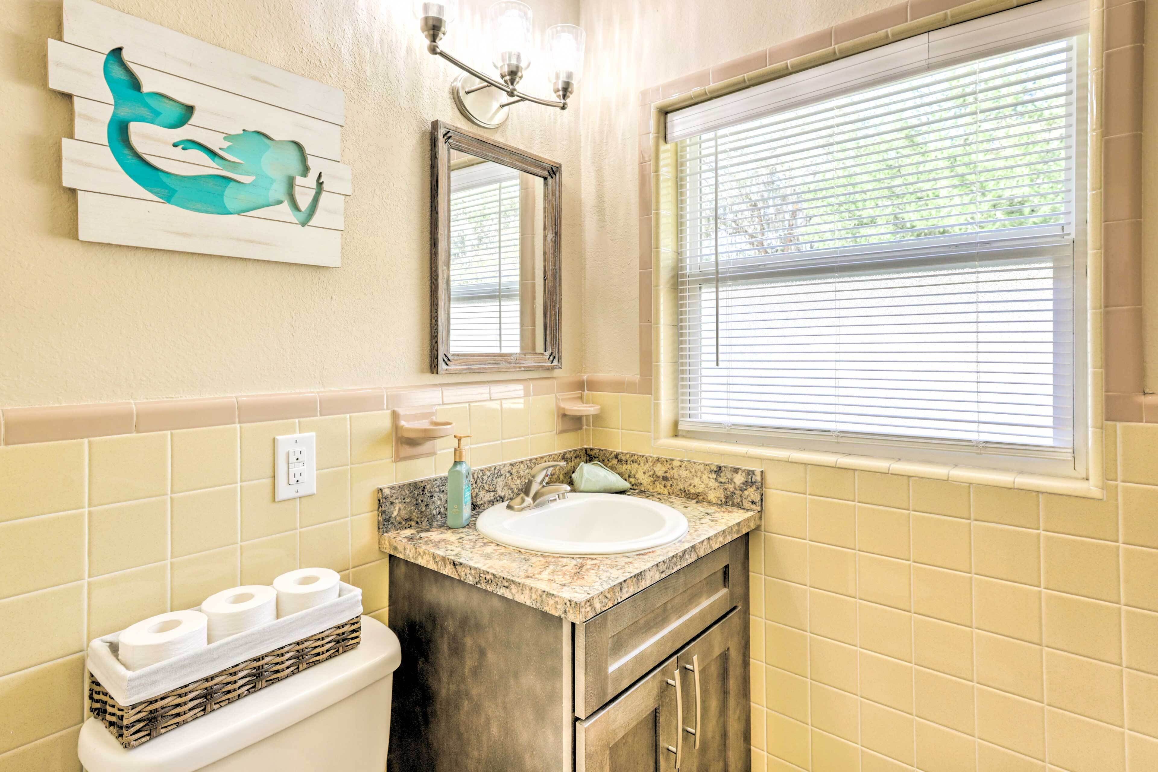 A bathroom with a sink, mirror, and toilet. There is a window with blinds and a wall-mounted light fixture.