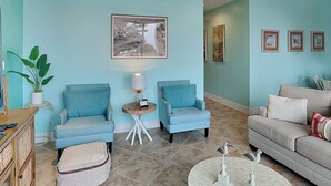 Una sala de estar con dos sillones azules, un sofá gris, una mesita redonda con una lámpara y un cuadro enmarcado en la pared.