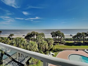 A balcony with a view of the beach, palm trees, and a pool.