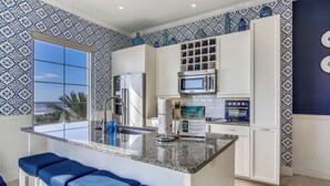 Una cucina con un'ampia finestra, mobili bianchi, un piano di lavoro in granito e sgabelli da bar blu.