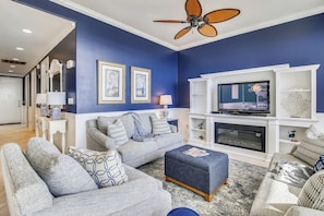 A living room with a blue accent wall, a grey sofa set, a television on a white entertainment unit, and a ceiling fan. - Blue Haven by Ryson Vacation Rentals (Galveston)