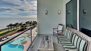 A balcony with a pool, a wooden table, a striped sofa, and a chair. - Precious Memories by Ryson Vacation Rentals (Galveston)