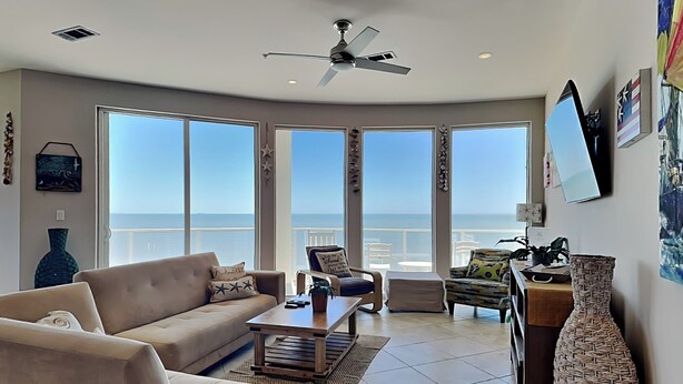 Een ruime woonkamer met een groot raam dat uitkijkt over de oceaan, een plafondventilator en uitzicht op de zee.