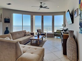 A spacious living room with a large window overlooking the ocean, a ceiling fan, and a view of the sea.