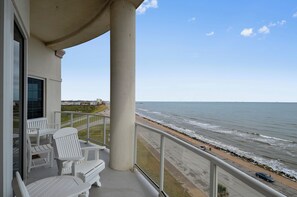 A balcony with white Adirondack chairs overlooking a beach with a rocky shoreline and a view of the ocean.