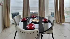 Un comedor con una mesa preparada para cuatro, ventanas grandes con cortinas y vista al océano.