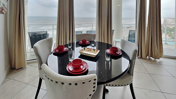 A dining area with a table set for four, large windows with curtains, and a view of the ocean.