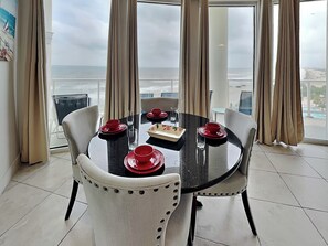 Un comedor con una mesa preparada para cuatro, ventanas grandes con cortinas y vista al océano.