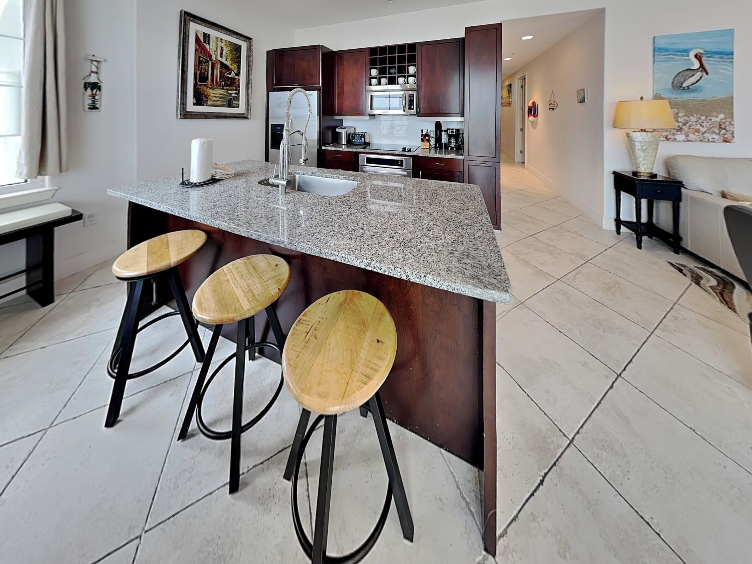 A kitchen with a granite countertop, wooden stools, dark cabinetry, and a view into the living area.