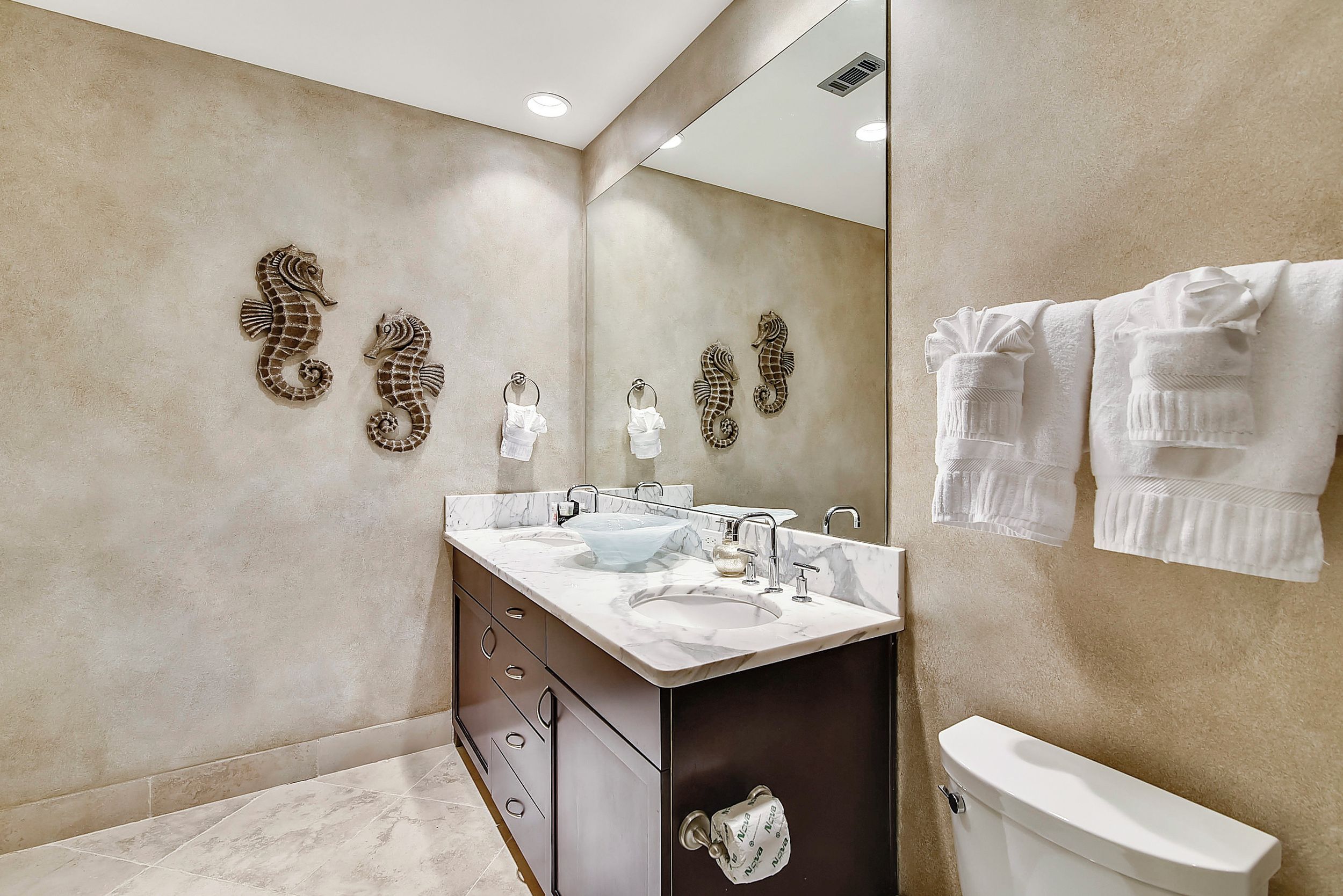A bathroom with a marble vanity, two sinks, a toilet, and decorative seahorse wall art.