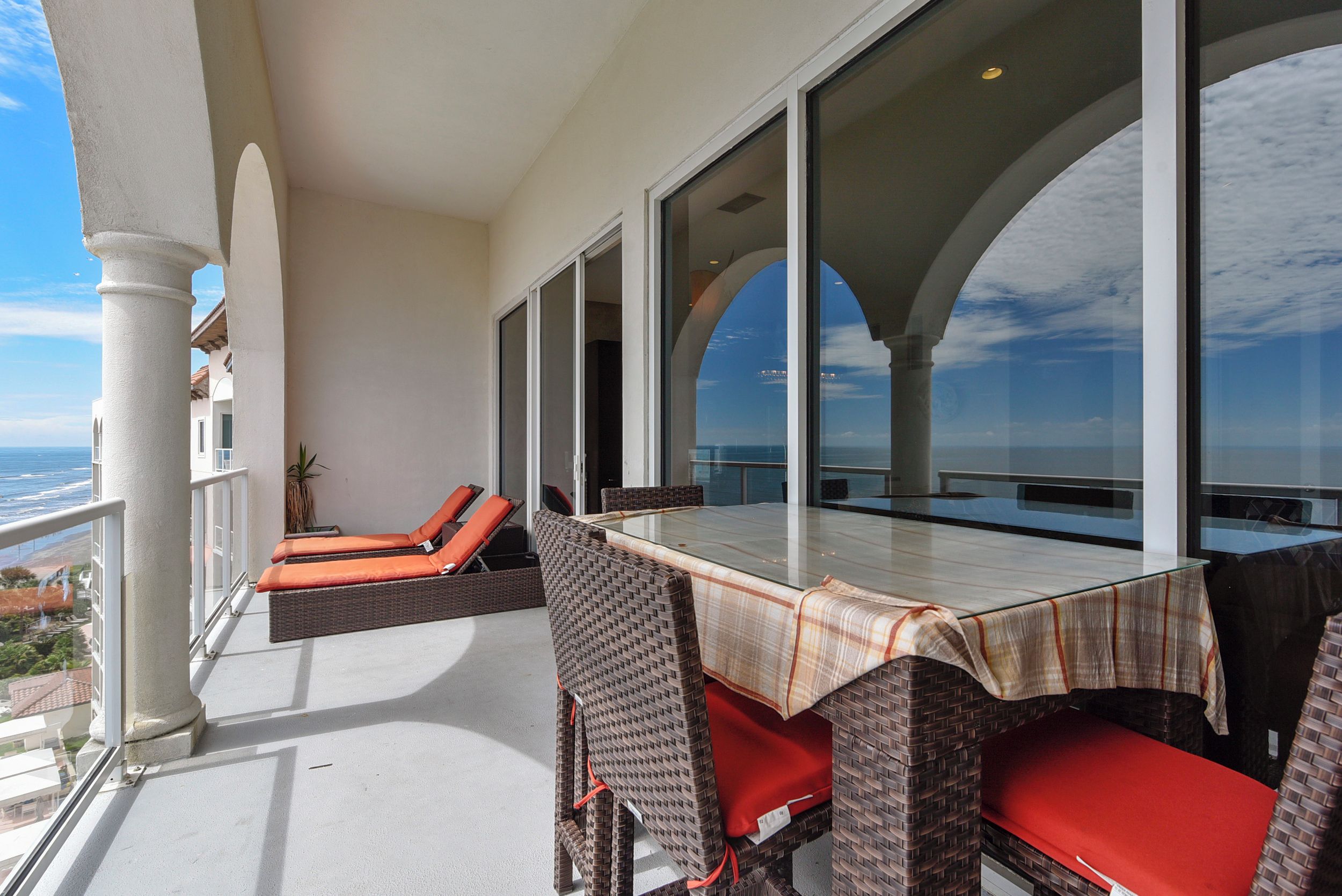 A balcony with wicker furniture and a glass table, overlooking a coastal view.