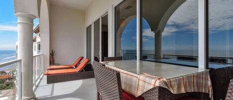 A balcony with wicker furniture and a glass table, overlooking a coastal view.
