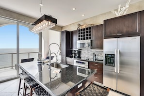 A modern kitchen with a large island, stainless steel appliances, and a view to the ocean. - Kohinoor by Ryson Vacation Rentals (Galveston)