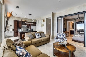 A modern living room with a sofa, armchair, and coffee table. A kitchen with a microwave and cabinets is visible in the background. - Kohinoor by Ryson Vacation Rentals (Galveston)