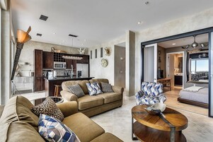 A modern living room with a sofa, armchair, and coffee table. A kitchen with a microwave and cabinets is visible in the background.