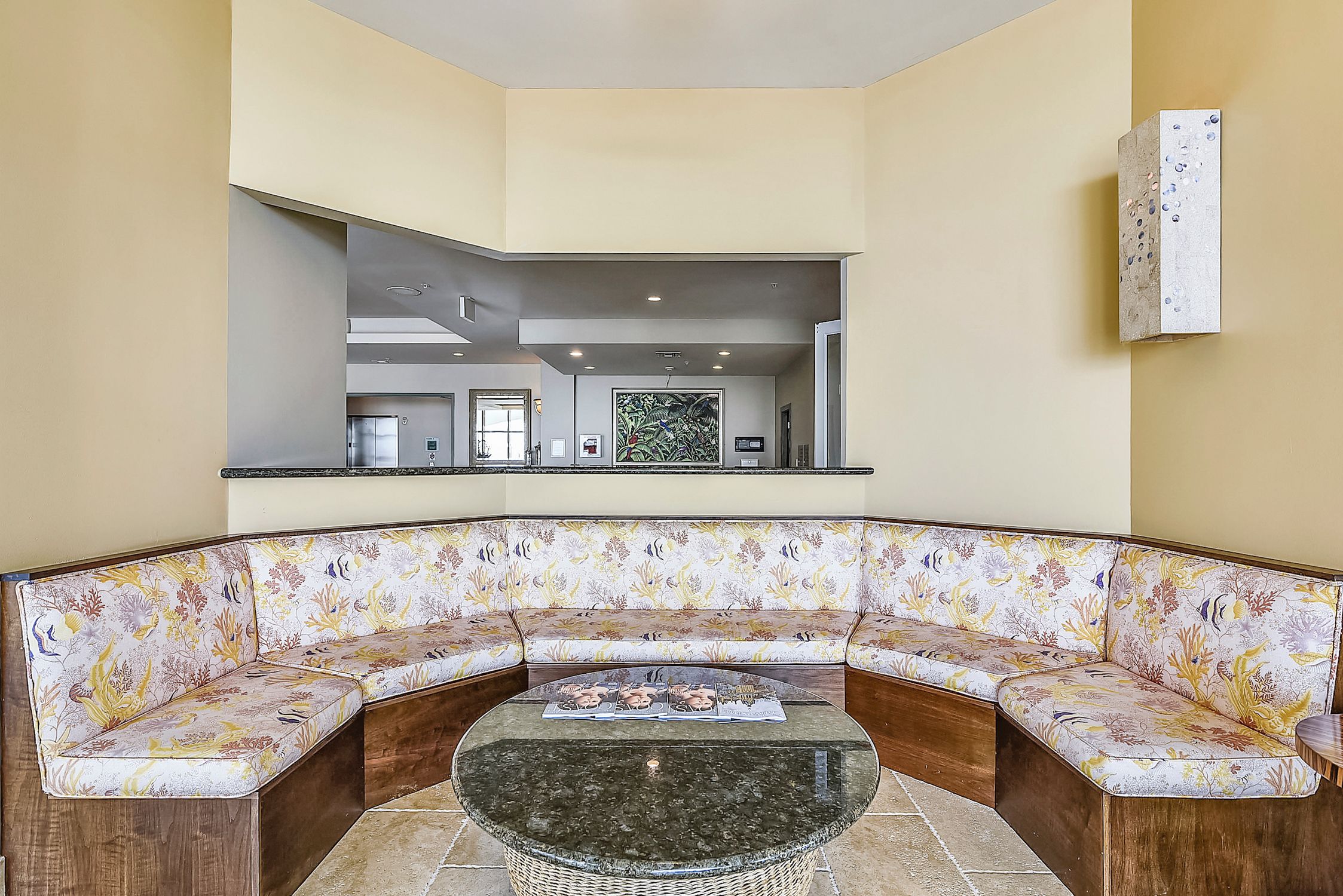 A corner seating area with a curved sofa and a round table, a wall-mounted artwork, and a view into the adjacent room.
