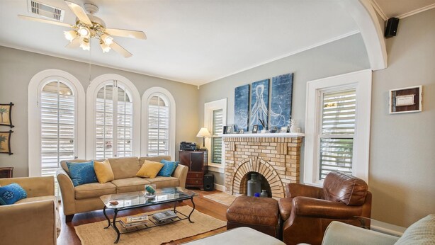 A living room with a fireplace, a ceiling fan, and a large window with shutters. - Endless Summer by Ryson Vacation Rentals (Galveston)