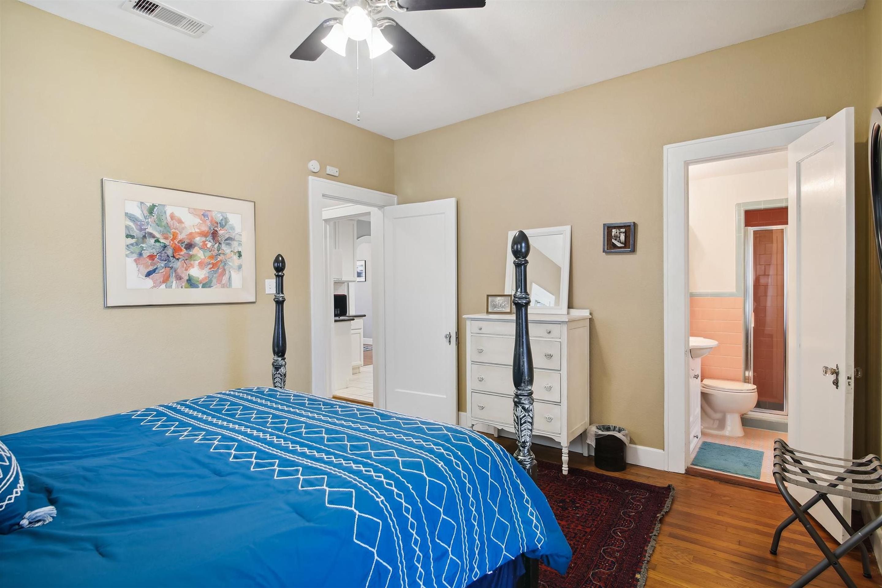 Quarto com cama, cômoda, mesa de cabeceira, ventilador de teto e porta do banheiro.
