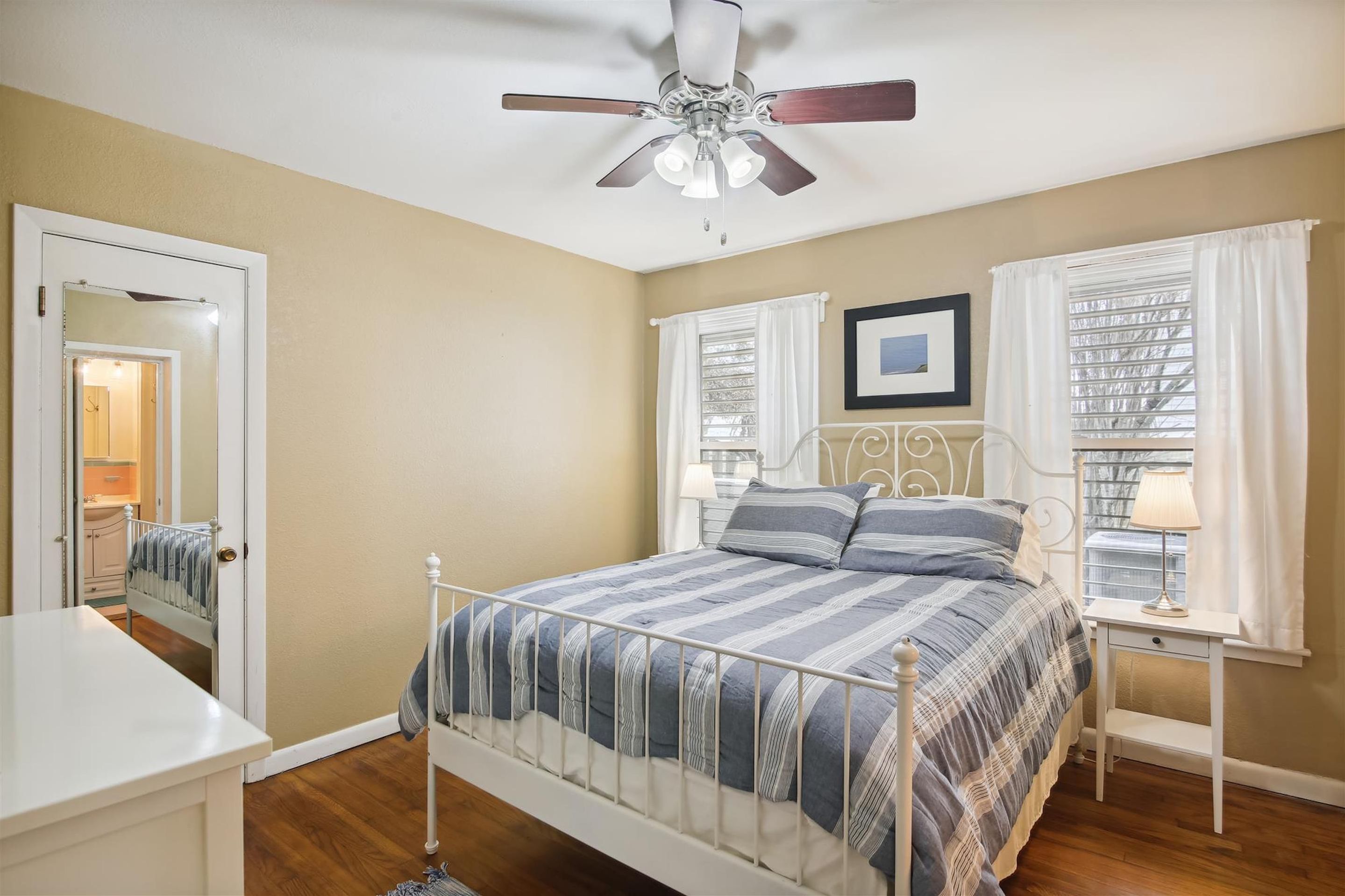 Une chambre à coucher comprenant un lit, un ventilateur de plafond, une table de chevet avec une lampe, une fenêtre avec des rideaux et une porte donnant sur une autre pièce.