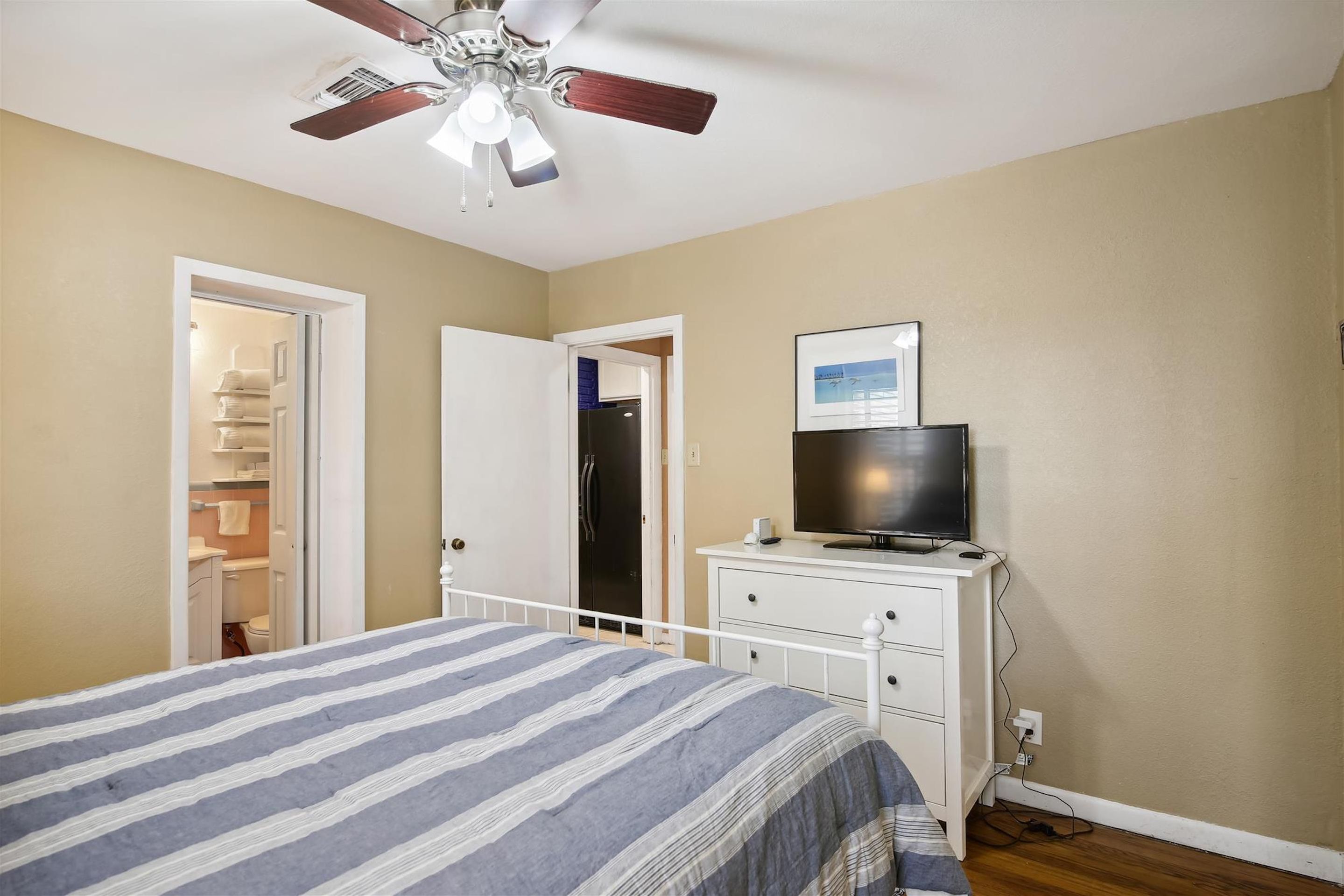 Une chambre à coucher avec un lit, un ventilateur de plafond, un téléviseur posé sur une commode et un placard avec un miroir.