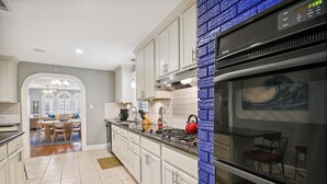 A kitchen with white cabinets, a blue brick accent wall, a stainless steel oven, and a dining area with a table and chairs. - Endless Summer by Ryson Vacation Rentals (Galveston)