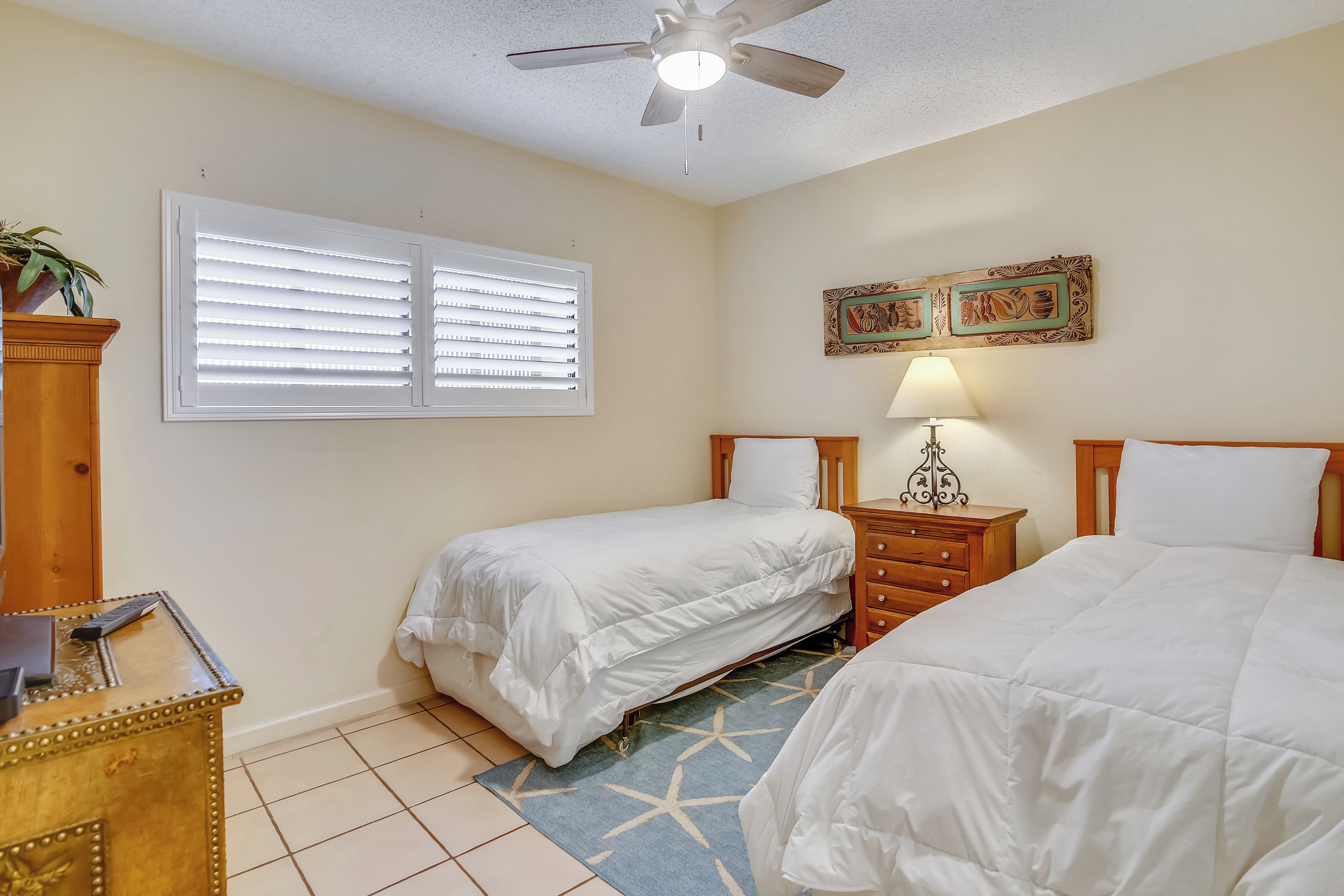 A bedroom with two beds, a ceiling fan, a window with blinds, a nightstand, a lamp, and a decorative wall piece.