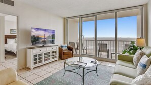 Una habitación de hotel con un balcón grande, televisión, sofá, mesa de centro y zona de comedor.