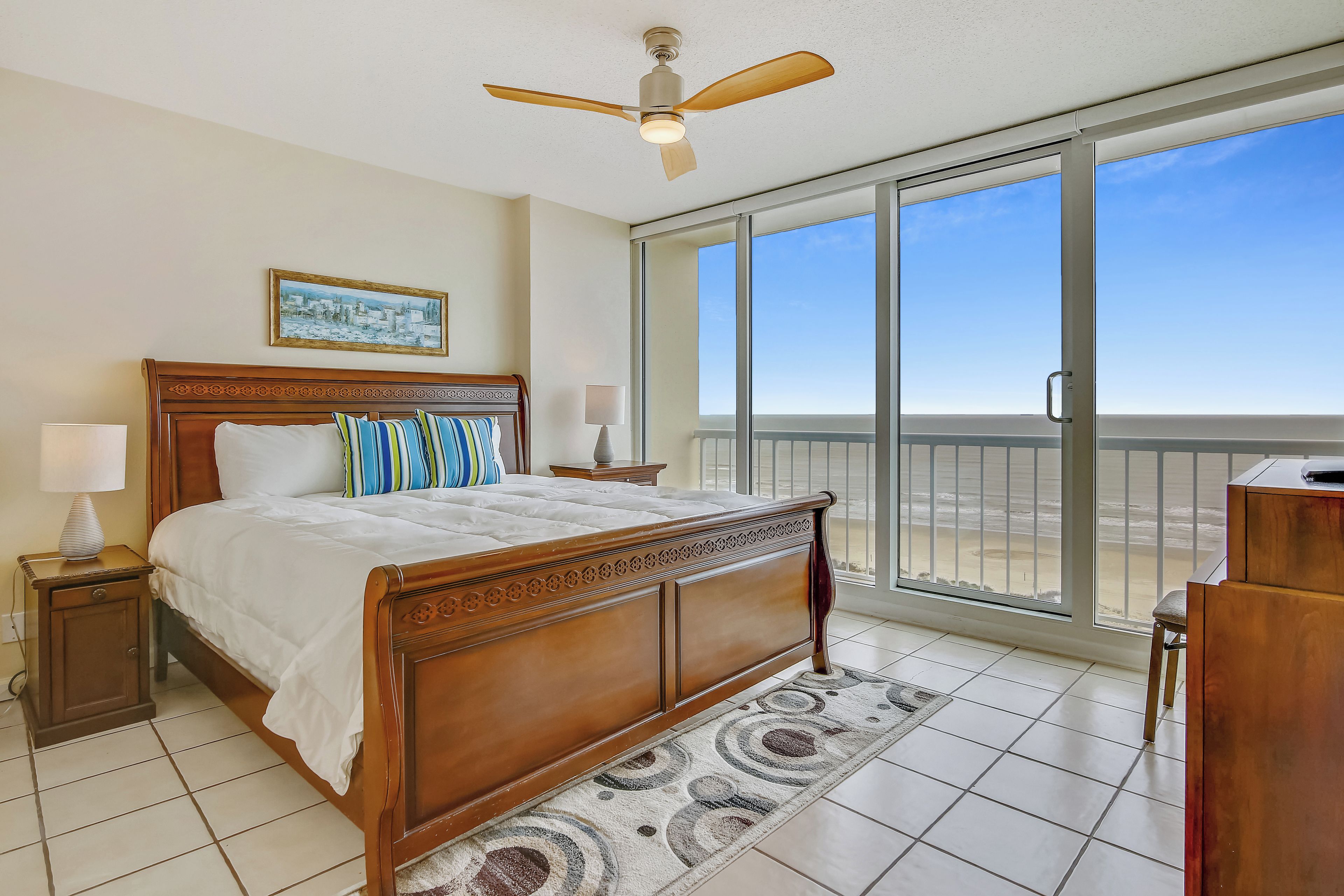 A bedroom with a large bed, a ceiling fan, a view of the beach, and a balcony.