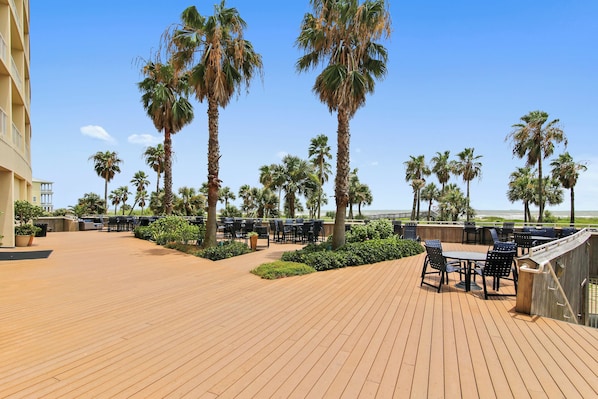 A wooden deck with outdoor seating, palm trees, and a view of the beach.