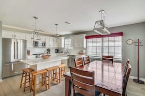 A modern kitchen with white cabinets, stainless steel appliances, and a wooden dining table with bar stools. - Herons Nest by Ryson Vacation Rentals (Galveston)