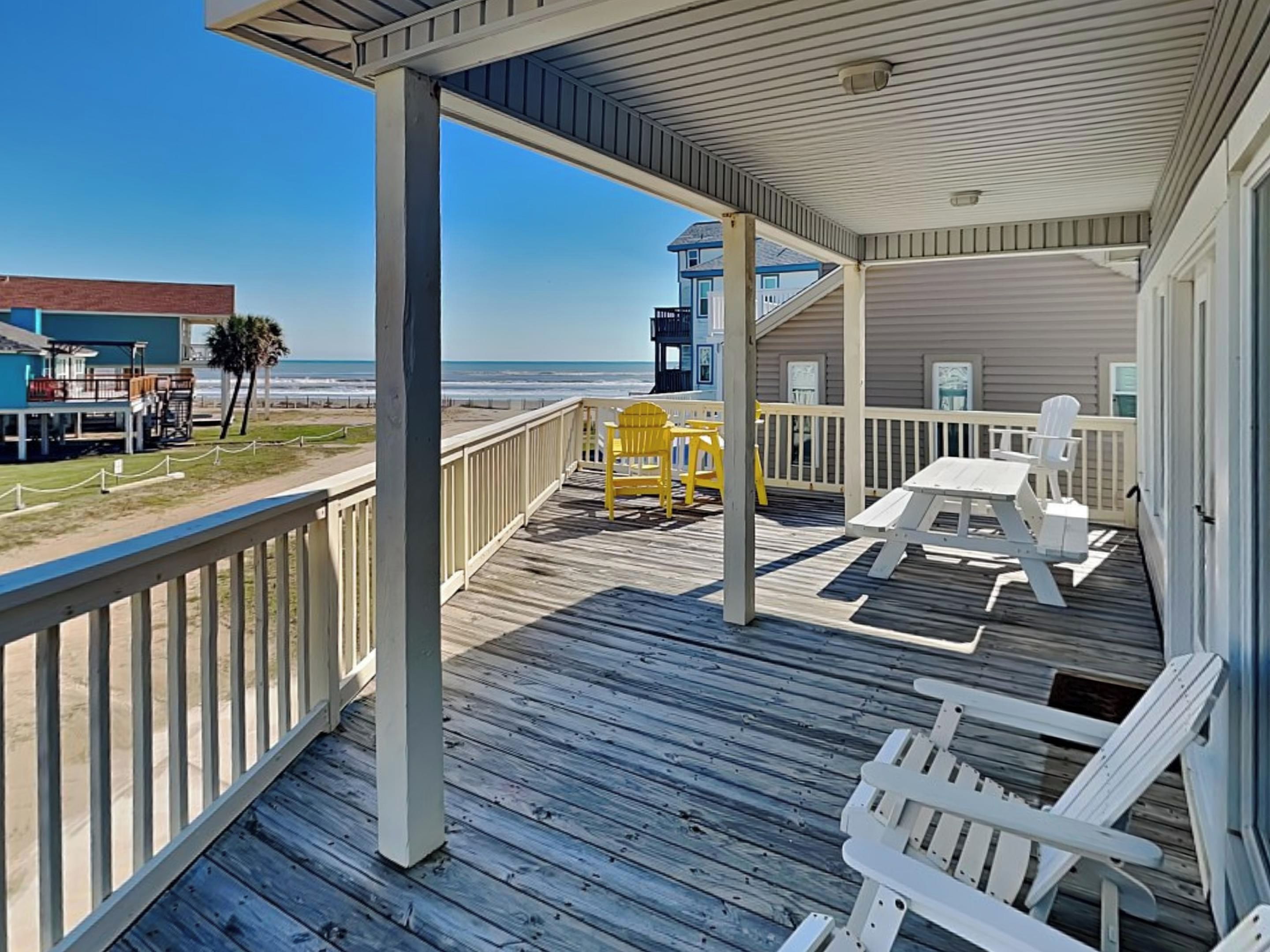 A wooden deck with white and yellow chairs, a table, and a view of the ocean and beach buildings.