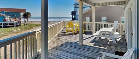 A wooden deck with white and yellow chairs, a table, and a view of the ocean and beach buildings.