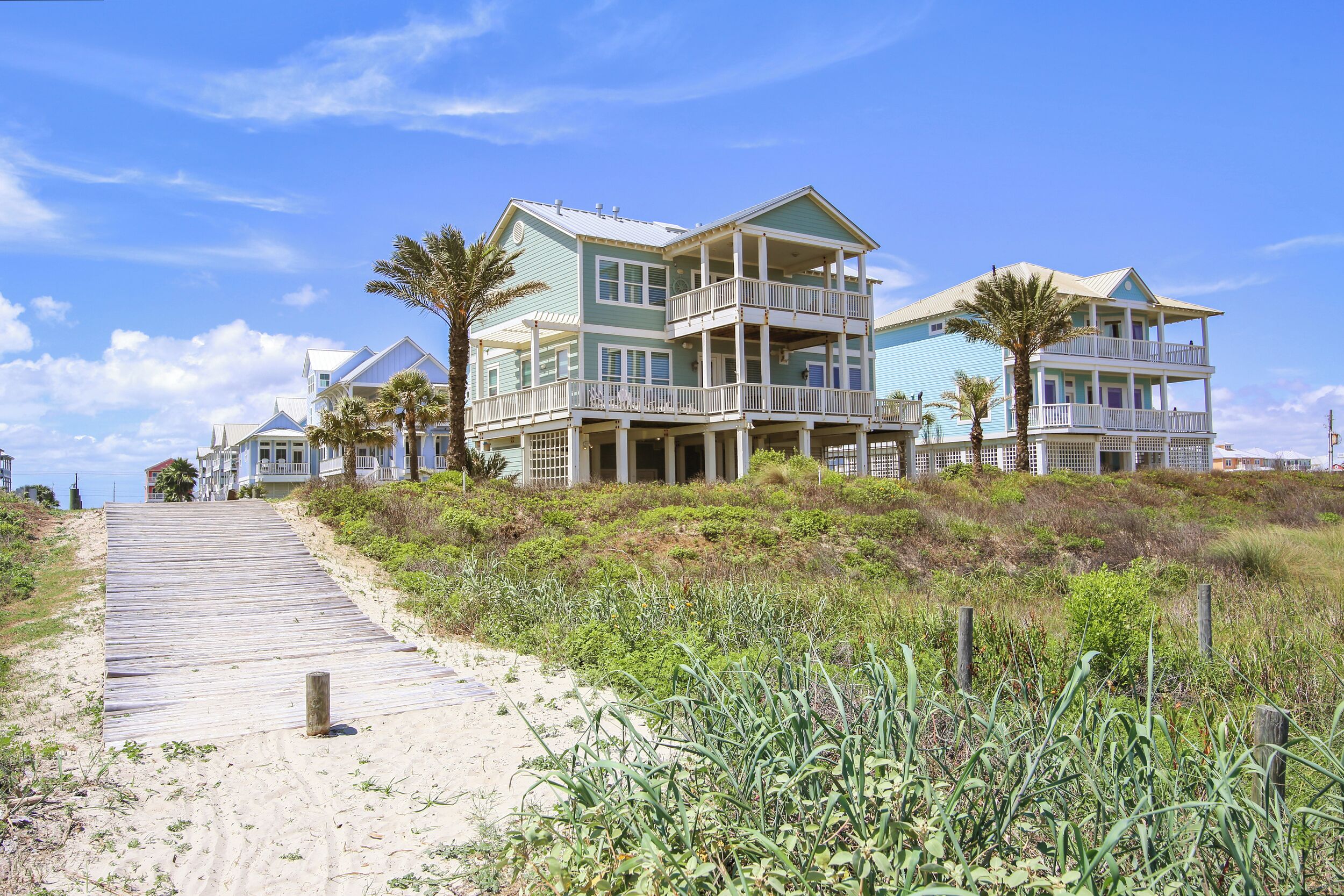 A beachfront property with a wooden boardwalk leading to a multi-story house with balconies, surrounded by vegetation and palm trees.