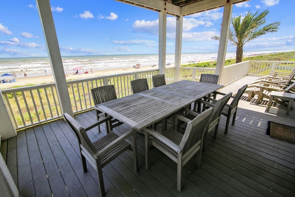 A wooden deck with a table and chairs, overlooking a beach with people and a palm tree.