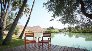 Een houten terras met twee houten stoelen gericht op een rustig meer, omgeven door palmbomen en begroeiing.