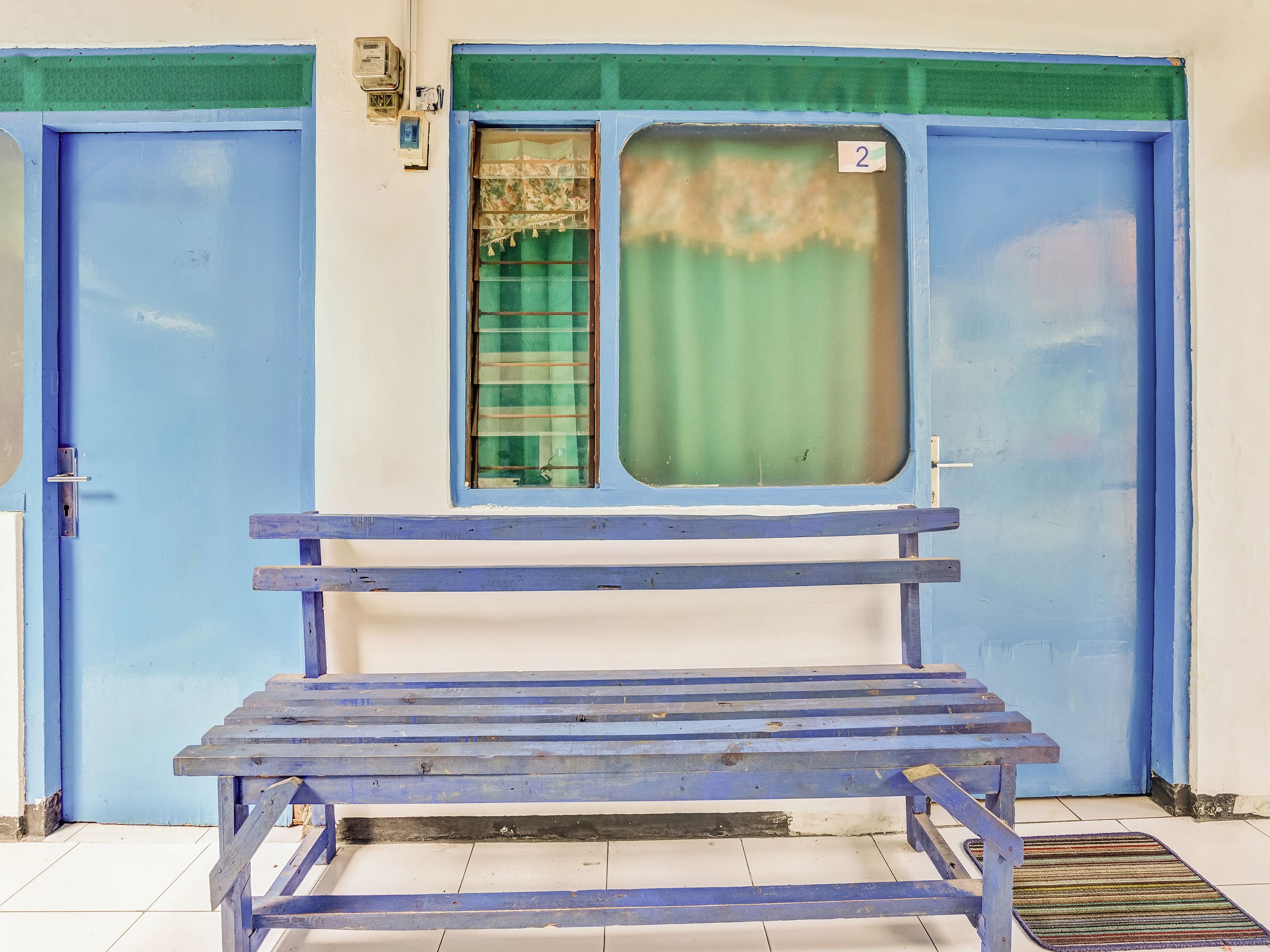 A blue bench in front of a building with two blue doors and a window with a green curtain.