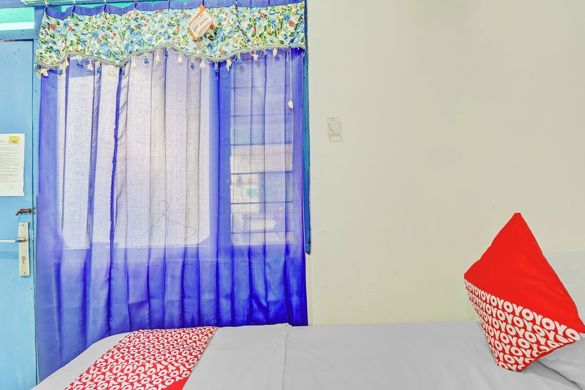 Ein Zimmer mit einem Bett, einem Fenster mit blauen Vorhängen, einem roten Kopfkissen und einer Tür mit blauem Rahmen.